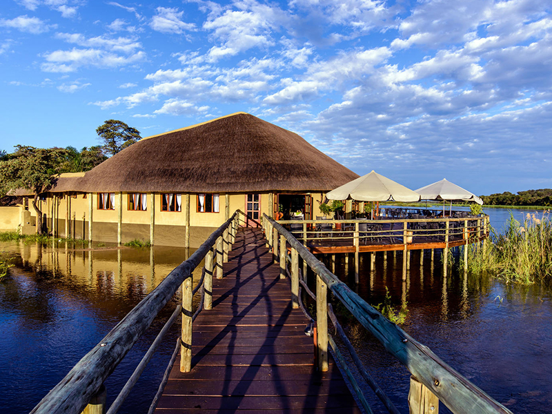 Wooden walkway leading to a large round thatched lodge building beside calm river water under a cloud-streaked sky, Namibia