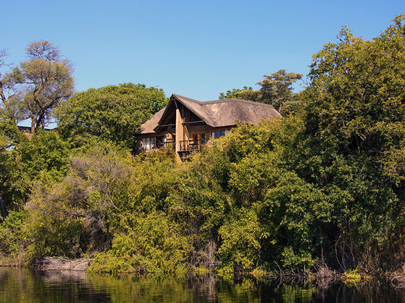 A thatched river lodge chalet set among dense green riverside trees under a clear blue sky, Zambezi Region, Namibia