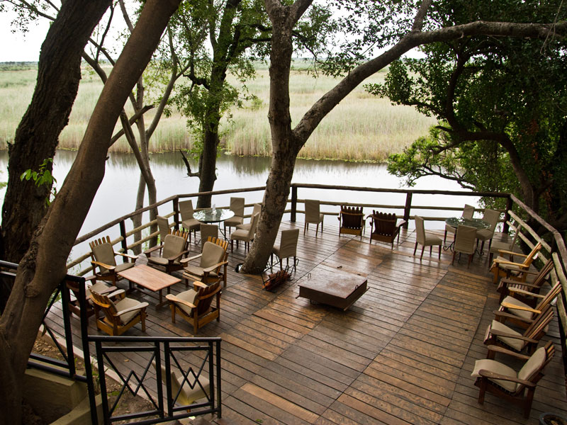 Wooden riverside deck with safari chairs and low tables under shade trees overlooking a reed-lined river, Zambezi Region, Namibia