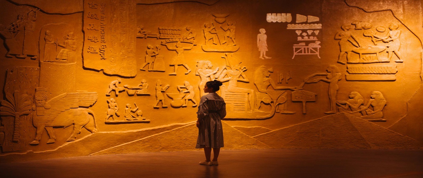 A visitor standing before a warmly lit wall of Egyptian-style carved reliefs in an indoor heritage exhibit
