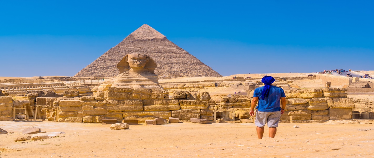 A visitor in a blue head covering walking across sand towards the Great Sphinx and a pyramid at Giza, Egypt