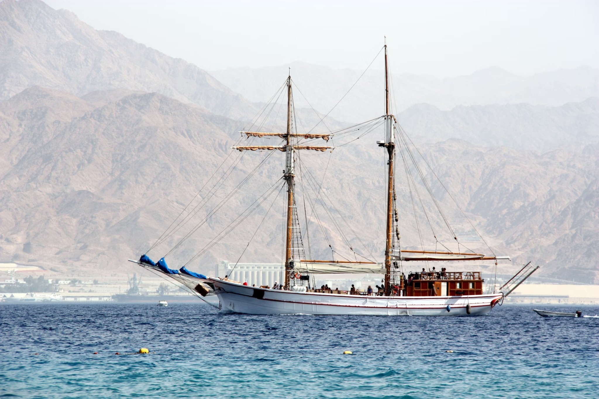 A traditional-style two-masted wooden sailing vessel anchored on calm blue water with arid mountains behind, Egypt